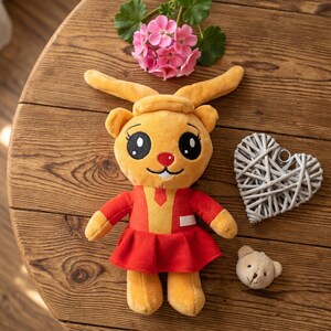 Puede incluir: Un peluche de conejito con grandes ojos negros, una nariz roja y un vestido rojo. El conejito es amarillo y naranja, con orejas largas y un sombrero pequeño. Una pequeña decoración en forma de corazón blanco y un pequeño osito de peluche están al lado del conejito.