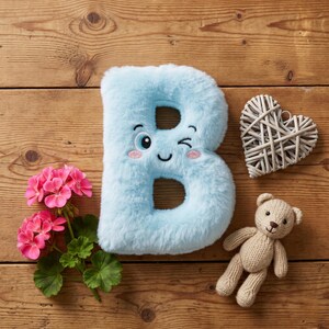 Puede incluir: Una letra "B" de felpa azul claro con una cara guiñando un ojo, un corazón de mimbre, un pequeño osito de peluche tejido y flores rosas sobre una superficie de madera. La letra tiene detalles bordados.