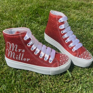 May include: A pair of high-top sneakers covered in red glitter with white rubber soles and white ribbon laces. The side of one sneaker features the text Mrs. Miller in silver glitter script. The toe caps are decorated with clear rhinestones.