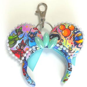 May include: A keychain featuring miniature mouse ears made of fabric with a colorful cartoon character print. The ears are trimmed with small white faux pearls and feature three light green oval gems in the center, attached to a silver metal clasp.