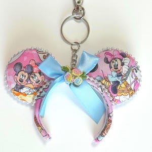May include: A keychain featuring two circular fabric ears with a pink background and Disney character prints, including Mickey and Minnie Mouse. The ears are accented with white faux pearls, a light blue satin bow, and a small floral embroidery.