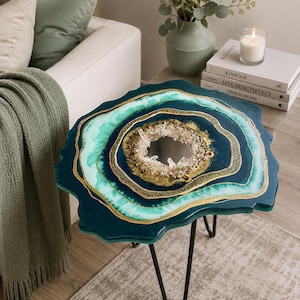 May include: A unique, handcrafted side table with a geode-inspired design. The table features a dark teal resin top with layers of teal and gold accents, and a central crystal cluster. It stands on black hairpin legs.