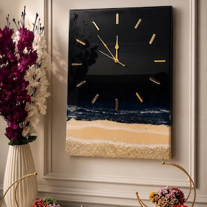 May include: A rectangular wall clock with a glossy black upper section and a textured beach scene at the bottom featuring sand and ocean waves. The clock face has gold-colored hour markers and matching gold-colored clock hands centered on the black area.