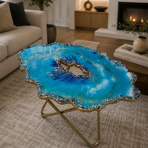Op de afbeelding: Een unieke, handgemaakte salontafel met een blauw en goud harsblad. De tafel heeft een vrije vorm met een goudkleurig metalen onderstel. De bovenkant heeft tinten blauw en goud, die op een agaatsteen lijken. De tafel is ongeveer 91 cm lang.