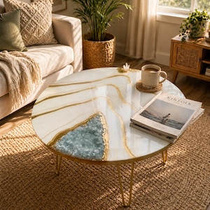 Könnte beinhalten: Ein runder Couchtisch mit einer weißen und goldenen Harzoberfläche, die ein geodenartiges Design mit einem blau-grünen Zentrum aufweist. Der Tisch hat goldfarbene Haarnadelbeine aus Metall und steht auf einem gewebten Teppich. Eine Tasse, ein Untersetzer und eine Zeitschrift liegen auf dem Tisch.