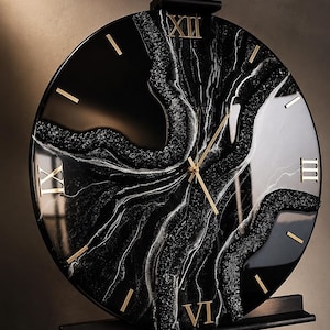 Può includere: Un orologio da parete circolare caratterizzato da una finitura in resina marmorizzata nera e bianca lucida con accenti glitterati testurizzati. Il quadrante mostra numeri romani dorati alle ore dodici, tre, sei e nove, con lancette dorate.