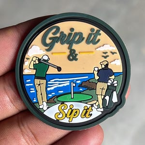 May include: A circular rubber patch featuring a golf scene with the text Grip it & Sip it. One figure swings a golf club while another stands with a drink and golf bag. The design includes a green, blue ocean, tan sky, and white clouds.
