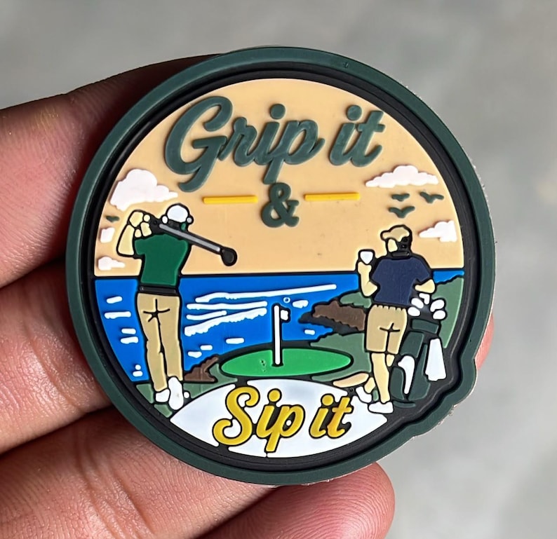 May include: A circular rubber patch featuring a golf scene with the text Grip it & Sip it. One figure swings a golf club while another stands with a drink and golf bag. The design includes a green, blue ocean, tan sky, and white clouds.