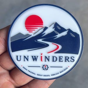 May include: A circular patch with a white background featuring a dark blue mountain range, a red sun, and a winding road. The text UNWINDERS is printed in dark blue with a red letter I. Below, the text reads GOOD FRIENDS, GREAT ROADS, ENDLESS HORIZONS.