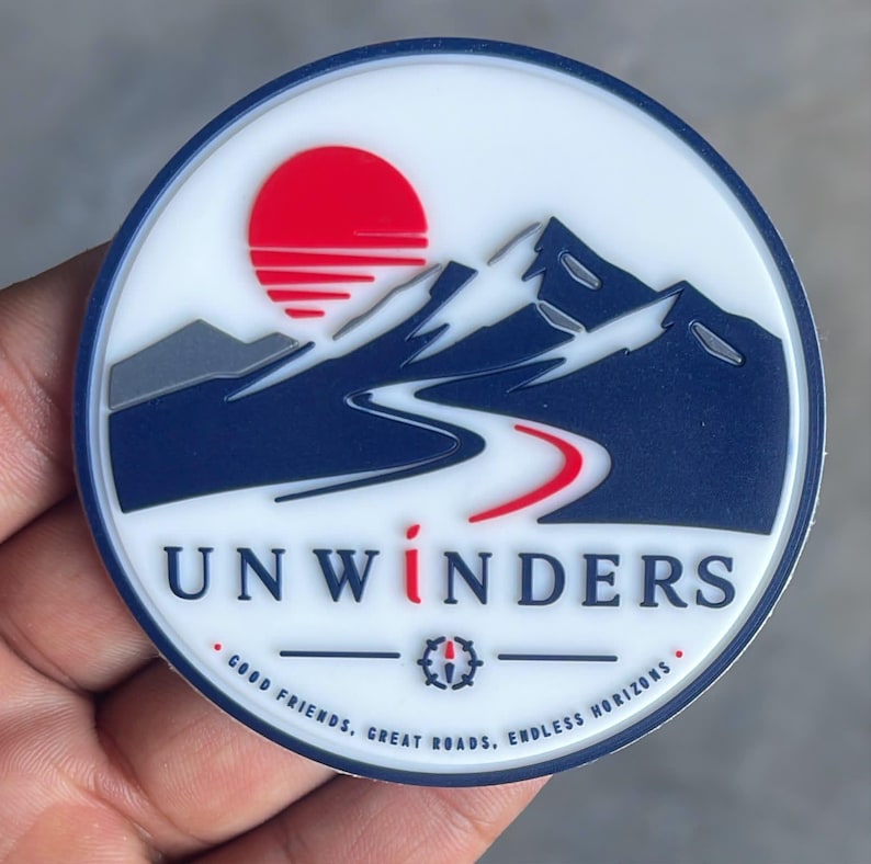 May include: A circular patch with a white background featuring a dark blue mountain range, a red sun, and a winding road. The text UNWINDERS is printed in dark blue with a red letter I. Below, the text reads GOOD FRIENDS, GREAT ROADS, ENDLESS HORIZONS.