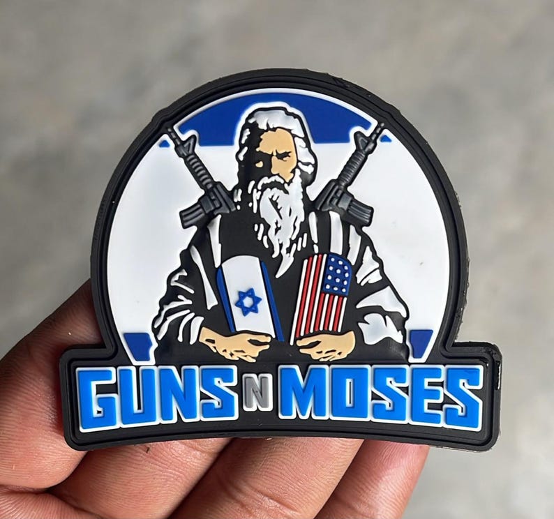 May include: A semi-circular PVC patch featuring a bearded figure holding an Israeli flag and an American flag, with two rifles crossed behind them. The text GUNS N MOSES is printed in bold blue letters at the bottom against a black border.