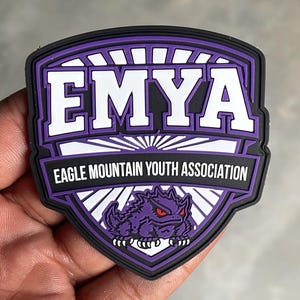 May include: A shield-shaped patch in purple, white, and black. The text EMYA appears in large white letters above the text EAGLE MOUNTAIN YOUTH ASSOCIATION. A graphic of a purple horned lizard with red eyes is positioned at the bottom of the patch.