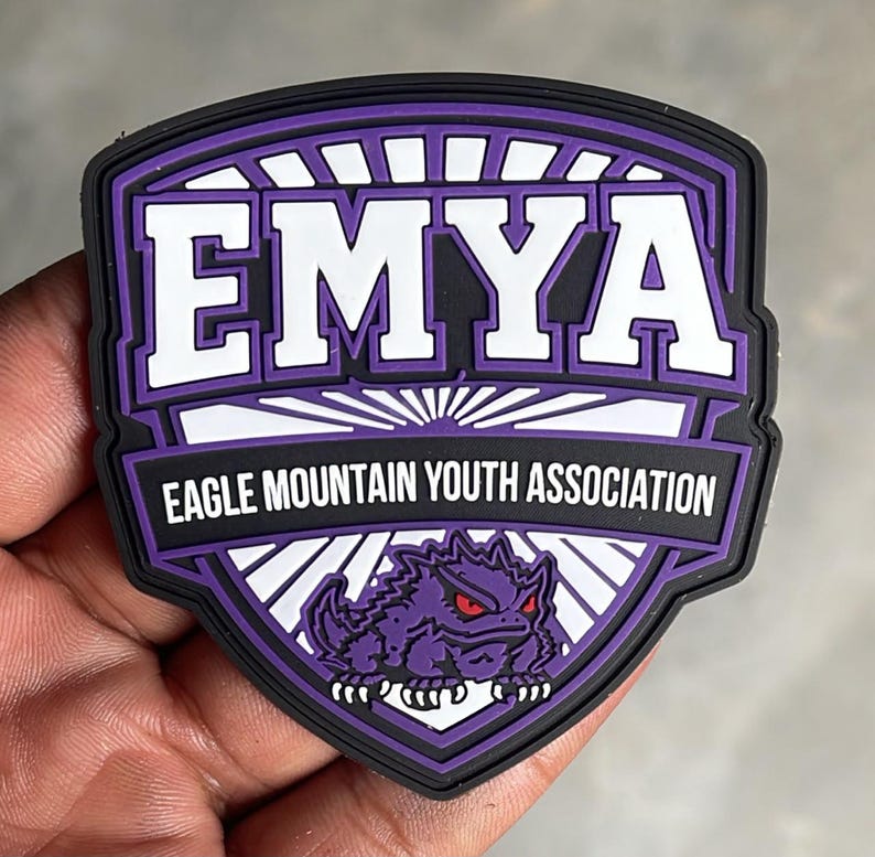 May include: A shield-shaped patch in purple, white, and black. The text EMYA appears in large white letters above the text EAGLE MOUNTAIN YOUTH ASSOCIATION. A graphic of a purple horned lizard with red eyes is positioned at the bottom of the patch.