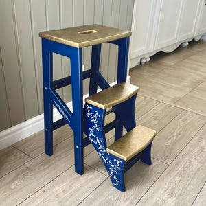 Puede incluir: Un taburete de madera de tres peldaños con una estructura de color azul intenso y patrones de volutas florales blancas en los paneles laterales. La superficie superior y los dos peldaños inferiores tienen un acabado en tono dorado.