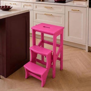 May include: A bright pink wooden step stool featuring three tiers and a rectangular top step with a cutout handle. The stool has a sturdy frame with side supports and a smooth finish, standing on a light-colored wooden floor in a kitchen setting.