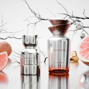Może przedstawiać: Dwa srebrne flakony perfum z brązowymi akcentami i napisami "MYKONOS" i "REFLECTION MYKONOS". Jeden flakon jest oznaczony "100ml 3.4 fl oz". Butelki znajdują się obok grejpfruta i dekoracyjnych gałęzi.