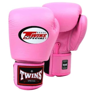 Puede incluir: Un par de guantes de boxeo de color rosa brillante con un parche ovalado blanco en la parte frontal que muestra el texto rojo Twins Special y cinco estrellas negras. La correa de la muñeca tiene una etiqueta rectangular negra con texto blanco.