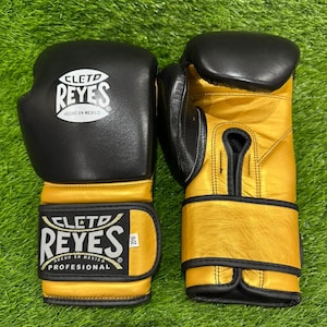 以下が含まれることがあります： 黒と金のボクシンググローブのペア。グローブには、ブランド名「CLETO REYES」が白い文字で表示されています。メキシコ製で、緑色の背景にあります。