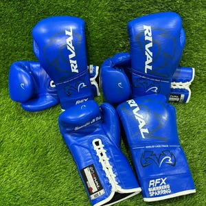Könnte beinhalten: Blaue Boxhandschuhe mit weißen Schnürsenkeln und dem Markennamen "RIVAL" aufgedruckt. Die Handschuhe liegen auf einer grünen Oberfläche. Der Text "RFX GUERRERO SPARRING" ist auf einem Handschuh zu sehen.