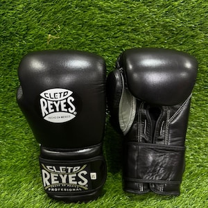 以下が含まれることがあります： 黒い革製ボクシンググローブのペアで、ブランド名「CLETO REYES」が白い文字で書かれています。グローブは緑の草の表面にあります。グローブはプロ仕様で、メキシコ製です。