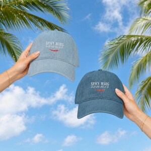 May include: Two baseball caps in light blue and dark blue denim fabric. Each cap features white embroidered text that reads SPICY MARG SUMMER with a small red chili pepper graphic centered below the text on the front panel of the hat.