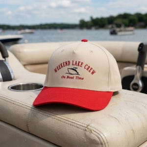May include: A beige and red baseball cap with the text "WEEKEND LAKE CREW" and "On Boat Time" in red. The cap features a boat graphic and a red brim. The cap is sitting on a boat seat with a lake in the background.