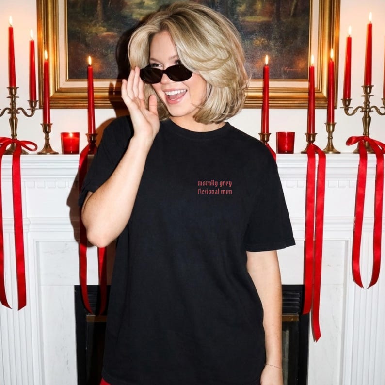 May include: Black t-shirt with the text "morally grey fictional men" in red script. The shirt is short-sleeved and worn by a person wearing sunglasses. Red candles and ribbons are in the background.