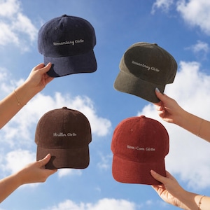 May include: Four corduroy baseball caps in navy blue, olive green, dark brown, and rust red. Each cap features embroidered white text on the front panel. The navy and olive caps read Romantasy Girlie, the brown cap reads Thriller Girlie, and the red cap reads Rom-Com Girlie.