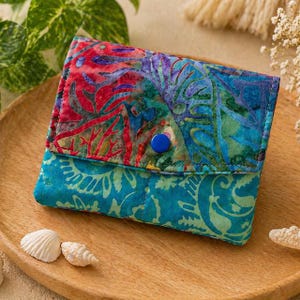May include: A colorful, quilted fabric wallet with a blue button closure. The top flap features a vibrant mix of red, blue, and purple patterns, while the bottom section is a teal and green design. The wallet is displayed on a wooden tray with seashells.