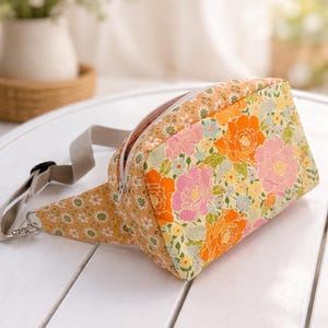 May include: A floral print fanny pack with a beige strap and zipper. The bag features a vibrant pattern of orange, pink, and yellow flowers on a light yellow background. The bag is lying on a white surface.