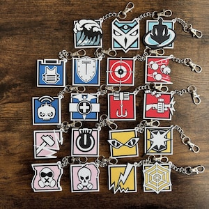 Puede incluir: Colección de llaveros cuadrados con varios diseños coloridos, incluyendo una ola, un escudo, una diana y una calavera. Cada llavero tiene una cadena y un cierre plateados. Los llaveros están dispuestos sobre una superficie de madera oscura.