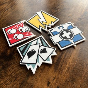 May include: Four square-shaped stickers featuring distinct graphic designs: a red icon with white figures, a yellow icon with a lightning bolt, a blue icon with a medical cross, and a light blue icon displaying two playing cards with spade symbols.