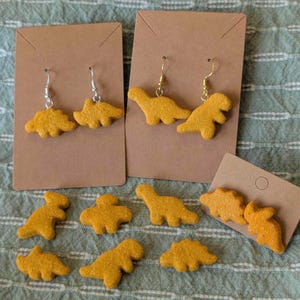 Op de afbeelding: Gele dinosaurussvormige oorbellen op bruine oorbellenkaarten. De oorbellen worden in paren getoond, met zilver- en goudkleurige haken. Extra dinosaurusvormen zijn verspreid rond de kaarten.