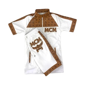 Puede incluir: Chándal blanco con detalles marrones y el logo MCM. La chaqueta tiene cuello, mangas y cremallera marrones. Los pantalones cortos a juego tienen el logo MCM en la pierna. El chándal está hecho de un material ligero.