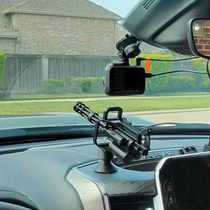 May include: A black miniature Gatling gun model mounted on a vehicle dashboard. The object features a rotating barrel assembly, a top handle, and a base stand. It is positioned near a digital dash camera attached to the windshield.