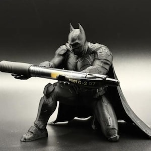 May include: A black Batman figurine in a kneeling position, holding a black Pilot G-2 07 gel pen horizontally with both hands. The figure features a textured suit, a cape extending behind it, and a pointed cowl. The pen has a clear barrel section.