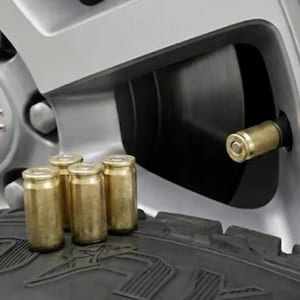 May include: A set of four brass-colored metal bullet-shaped tire valve stem caps. Three caps are resting on the black rubber tread of a tire, while one cap is installed on the valve stem of a silver GMC vehicle wheel with visible lug nuts.