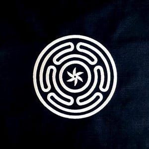 Puede incluir: Textil rectangular negro con un diseño circular blanco en el centro. El diseño presenta un patrón laberíntico que rodea una estrella de seis puntas. El textil es probablemente un artículo decorativo, como un mantel individual.