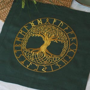 May include: A square dark green fabric napkin featuring a central gold embroidered design of a tree of life enclosed within a circular border of runic characters. The embroidery is detailed with intricate knotwork patterns throughout the tree.