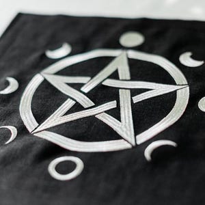 Puede incluir: Tela cuadrada negra con un diseño de pentagrama bordado en blanco. El pentagrama está rodeado por un círculo y formas de media luna. Un círculo y un punto están debajo.