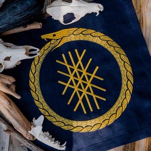 Peut inclure: Un textile bleu marine présente un motif d'ouroboros brodé en or, un serpent se mordant la queue, encerclant un motif géométrique doré. Le textile est entouré de crânes d'animaux et de branches en bois. L'ouroboros est détaillé avec des écailles.