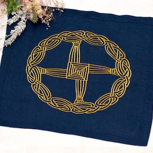 Puede incluir: Un mantel individual rectangular de tela azul marino con un diseño bordado en amarillo dorado centrado que muestra una cruz de Santa Brígida dentro de un patrón circular de nudos celtas. El tejido tiene una trama visible y un borde cosido.