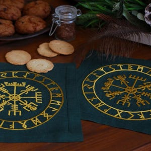 Peut inclure: Deux sets de table vert foncé avec des symboles vikings brodés en or. Les sets de table sont rectangulaires et présentent un motif central en forme de boussole entouré de runes. L'image comprend également des biscuits, des épices et des éléments décoratifs.
