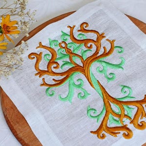Peut inclure: Serviette carrée en lin blanc avec un motif d'arbre brodé en marron et vert. La serviette est posée sur une surface en bois, avec des fleurs jaunes et du gypsophile blanc en arrière-plan.