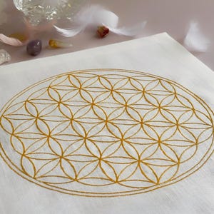 以下が含まれることがあります： 白い布の中央に、ゴールドカラーの刺繍でフラワー・オブ・ライフの模様が円形に施されています。この幾何学的なモチーフは、複数の円が重なり合い、対称的で繰り返しのある花のような格子状の構造を形成しています。