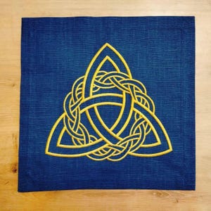 Könnte beinhalten: Ein quadratischer, marineblauer Stoff mit einem goldfarbenen keltischen Knotenmuster, das in der Mitte gestickt ist. Der Knoten ist eine komplexe, ineinander verschlungene dreieckige Form.