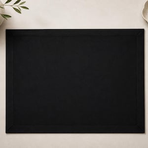 以下が含まれることがあります： 中央が単色で、幅の広いステッチ入りの縁取りが施された長方形の黒いランチョンマット。表面は平らで清潔感があり、角は額縁仕立てになっており、テーブルセッティングに整然とした統一感のある外観を与えています。