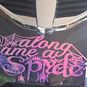 Könnte beinhalten: Ein rosa-lila Farbverlauf-Aufkleber mit den Worten "along came a spider" in einer stilisierten Schriftart, mit Spinnennetzen und Spinnenillustrationen. Der Aufkleber ist auf einer dunkelgrauen Oberfläche mit Chromakzent angebracht.