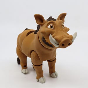 May include: A tan-colored, cartoon-style warthog figurine with large tusks and a smiling expression. The figurine has articulated joints and is posed on a white surface. The warthog has dark brown hair and light gray hooves.