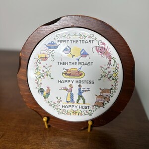 Puede incluir: Un salvamanteles redondo y vintage con un diseño bordado a punto de cruz. El centro presenta el texto "FIRST THE TOAST THEN THE ROAST" y "HAPPY HOSTESS HAPPY HOST". Está enmarcado en madera oscura y tiene un soporte amarillo.
